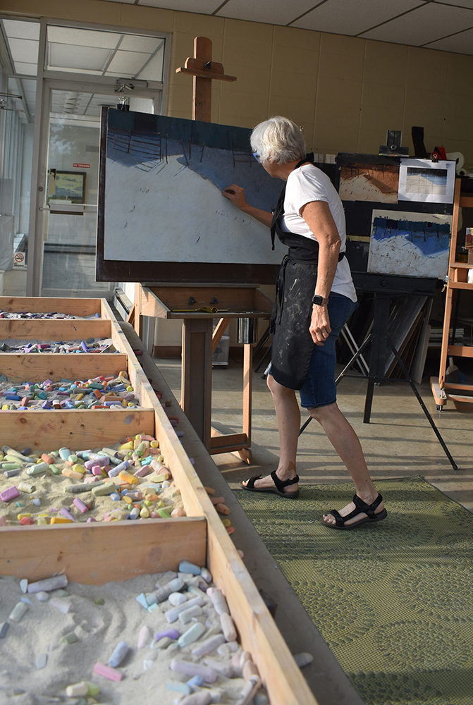 Woman artist painting on an easel in her studio with trays of pastels in the foreground