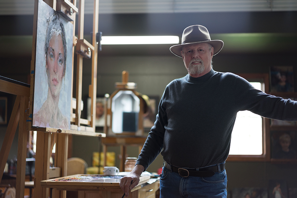 Male artist in his studio next to a painting on an easel