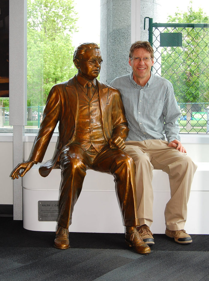 Male sculptor sitting next to a life size sculpture of a man in a suit