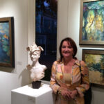 Artist Spotlight: Marcia Holmes photo of artist standing in front of two paintings, and next to a head bust of a woman