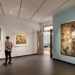 National Art Museum Reopens This Month A modern art museum gallery is photographed at a wide angle. A visitor observes framed artworks on the walls to the left. To the right of the visitor, a large mixed media painting shows a downtrodden woman walking with a hulking load of corn on her back. Several birds stand at her feet.