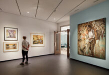 National Art Museum Reopens This Month A modern art museum gallery is photographed at a wide angle. A visitor observes framed artworks on the walls to the left. To the right of the visitor, a large mixed media painting shows a downtrodden woman walking with a hulking load of corn on her back. Several birds stand at her feet.