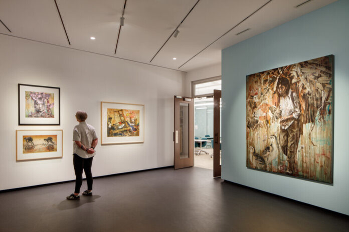 A modern art museum gallery is photographed at a wide angle. A visitor observes framed artworks on the walls to the left. To the right of the visitor, a large mixed media painting shows a downtrodden woman walking with a hulking load of corn on her back. Several birds stand at her feet.