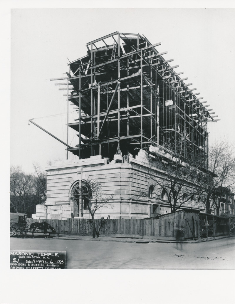 A black and white archival photo of NMWA's building during construction taken from the street. There is a wooden fence around the base of the building with a horsedrawn carriage parked on the street. A person, represented only as a blur, is crossing the street. Art museum construction.