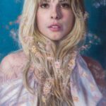 Featured Artwork: Nanci France-Vaz oil painting of woman looking at viewer, holding her long blonde hair; blue pattern background